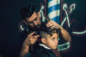 A bearded, tattooed barber cuts a young boy’s hair with clippers and a comb, as a barber pole and scissors decorate the background.